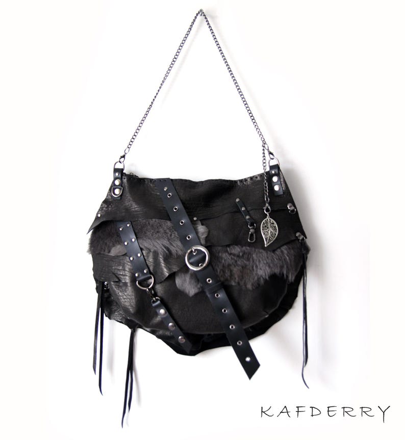 Puede incluir: Bolso de hombro de cuero negro con correa de cadena, con un panel de piel y correas decorativas con herrajes plateados. Un dije de hoja plateada cuelga de la cadena. El bolso tiene un dise&ntilde;o asim&eacute;trico &uacute;nico. La marca "KAFDERRY" est&aacute; impresa.