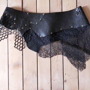May include: A black leather belt featuring a wide, contoured waistband adorned with metallic studs and a single D-ring. Attached to the lower edge are layered, asymmetrical panels of black mesh and intricate, web-like textured fabric.