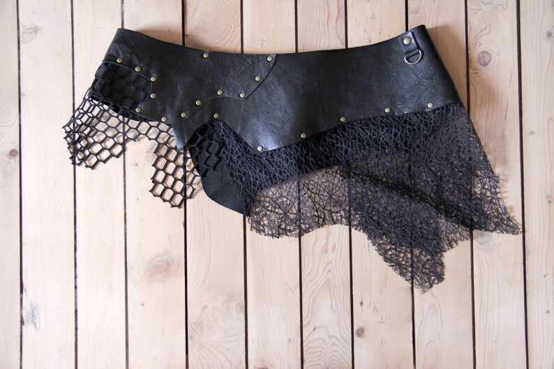 May include: A black leather belt featuring a wide, contoured waistband adorned with metallic studs and a single D-ring. Attached to the lower edge are layered, asymmetrical panels of black mesh and intricate, web-like textured fabric.