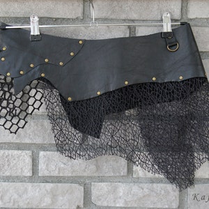 May include: A black skirt featuring a smooth leather-like upper panel adorned with small brass-colored metal studs and a circular ring attachment. The lower section consists of layered black mesh fabric with a grid pattern and an irregular hemline.