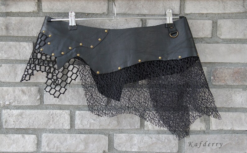 May include: A black skirt featuring a smooth leather-like upper panel adorned with small brass-colored metal studs and a circular ring attachment. The lower section consists of layered black mesh fabric with a grid pattern and an irregular hemline.