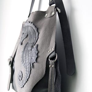 May include: Grey leather shoulder bag with a seahorse appliqu&eacute;. The bag features a dark grey seahorse design on the front, with silver rivets and black leather straps. The bag is hanging against a white wall.
