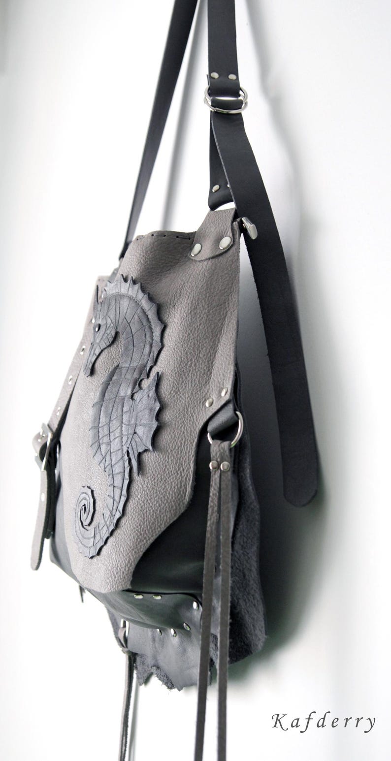 May include: Grey leather shoulder bag with a seahorse appliqu&eacute;. The bag features a dark grey seahorse design on the front, with silver rivets and black leather straps. The bag is hanging against a white wall.