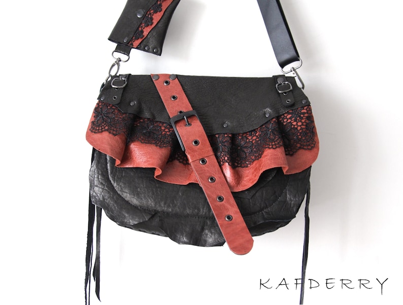 K&ouml;nnte beinhalten: Schwarze Leder-Umh&auml;ngetasche mit roter Lederklappe und schwarzem Spitzensaum. Die Tasche hat einen roten Lederriemen mit Schnalle und einen schwarzen Schulterriemen. Die Tasche ist mit Nieten verziert und tr&auml;gt das Wort "KAFDERRY".
