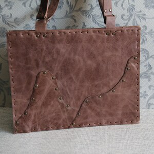 May include: A rectangular brown leather tote bag featuring visible hand-stitched edges and metallic rivets. The front panel displays an overlapping leather layer with a mountain-like silhouette, secured by small brass-toned metal studs.