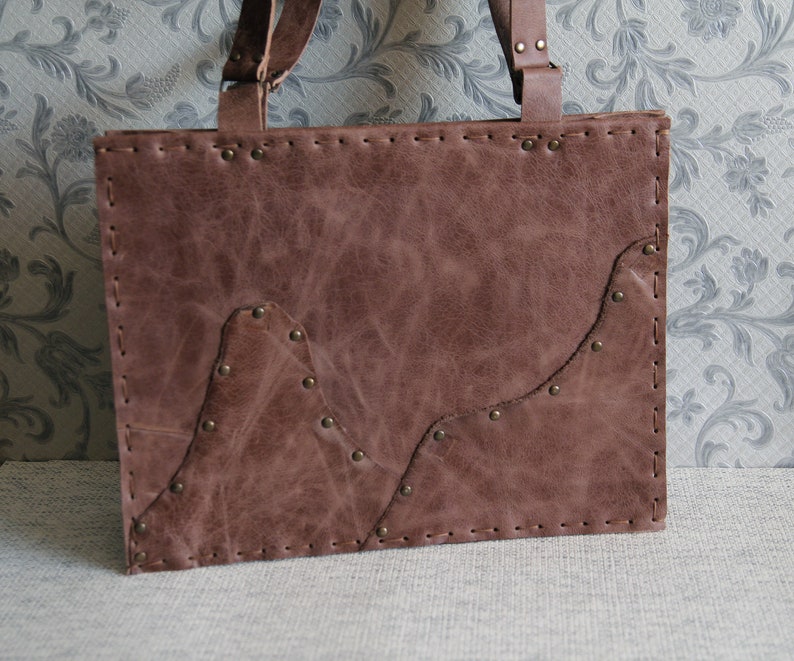 May include: A rectangular brown leather tote bag featuring visible hand-stitched edges and metallic rivets. The front panel displays an overlapping leather layer with a mountain-like silhouette, secured by small brass-toned metal studs.