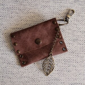 May include: A brown rectangular pouch with a textured surface, secured by a central snap button. The pouch features metallic rivets along the edges and is attached to a bronze-toned lobster clasp keychain with a dangling leaf-shaped metal charm.