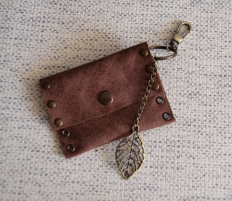 May include: A brown rectangular pouch with a textured surface, secured by a central snap button. The pouch features metallic rivets along the edges and is attached to a bronze-toned lobster clasp keychain with a dangling leaf-shaped metal charm.
