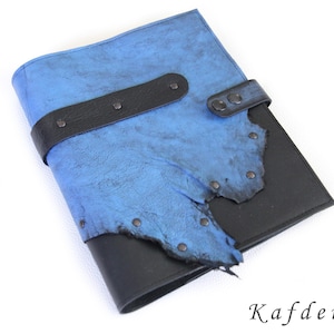 Puede incluir: Una cubierta de diario rectangular con una base negra lisa y una capa superior azul texturizada con bordes irregulares. Una correa negra con remaches metálicos y un cierre a presión asegura la parte frontal. La palabra Kafderry aparece impresa abajo.