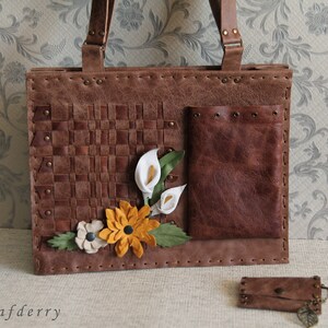 May include: A brown leather tote bag featuring a woven panel, a smooth pocket with metal grommets, and decorative leather flowers in white, yellow, and beige. The bag has two handles and is accompanied by a small matching brown leather coin purse.