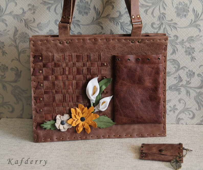 May include: A brown leather tote bag featuring a woven panel, a smooth pocket with metal grommets, and decorative leather flowers in white, yellow, and beige. The bag has two handles and is accompanied by a small matching brown leather coin purse.