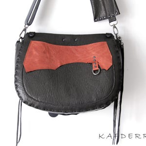 K&ouml;nnte beinhalten: Eine schwarze Leder-Umh&auml;ngetasche mit einem rostfarbenen Akzentpaneel. Die Tasche hat eine gebogene Form, einen langen Riemen und dekorative N&auml;hte. Die Tasche hat einen Metallring und Nieten. Das Wort "KAFDERRY" ist unten aufgedruckt.