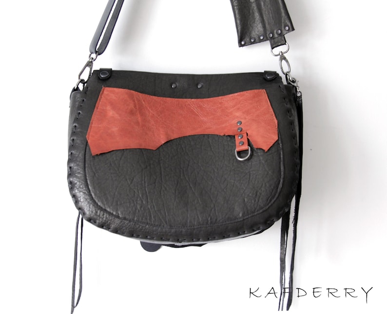 K&ouml;nnte beinhalten: Eine schwarze Leder-Umh&auml;ngetasche mit einem rostfarbenen Akzentpaneel. Die Tasche hat eine gebogene Form, einen langen Riemen und dekorative N&auml;hte. Die Tasche hat einen Metallring und Nieten. Das Wort "KAFDERRY" ist unten aufgedruckt.