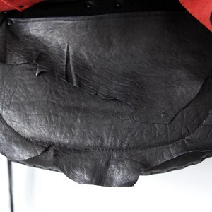 K&ouml;nnte beinhalten: Eine schwarze Ledertasche mit strukturierter Oberfl&auml;che und einem einzigartigen, geschichteten Design. Die Tasche hat einen Klappenverschluss und lange, d&uuml;nne Riemen. Der obere Teil der Tasche ist rot mit schwarzen Verzierungen.