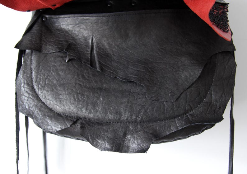 K&ouml;nnte beinhalten: Eine schwarze Ledertasche mit strukturierter Oberfl&auml;che und einem einzigartigen, geschichteten Design. Die Tasche hat einen Klappenverschluss und lange, d&uuml;nne Riemen. Der obere Teil der Tasche ist rot mit schwarzen Verzierungen.