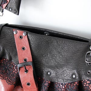 K&ouml;nnte beinhalten: Schwarze und rote Leder-Umh&auml;ngetasche mit dekorativer Spitzenborte. Die Tasche hat einen roten Riemen mit schwarzer Schnalle und silberfarbener Hardware. Das Design der Tasche umfasst Nieten und eine strukturierte Lederoberfl&auml;che.