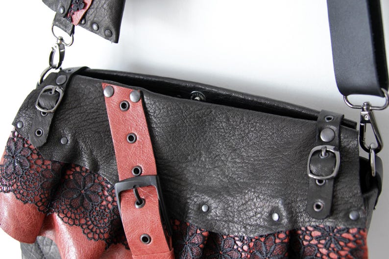 K&ouml;nnte beinhalten: Schwarze und rote Leder-Umh&auml;ngetasche mit dekorativer Spitzenborte. Die Tasche hat einen roten Riemen mit schwarzer Schnalle und silberfarbener Hardware. Das Design der Tasche umfasst Nieten und eine strukturierte Lederoberfl&auml;che.