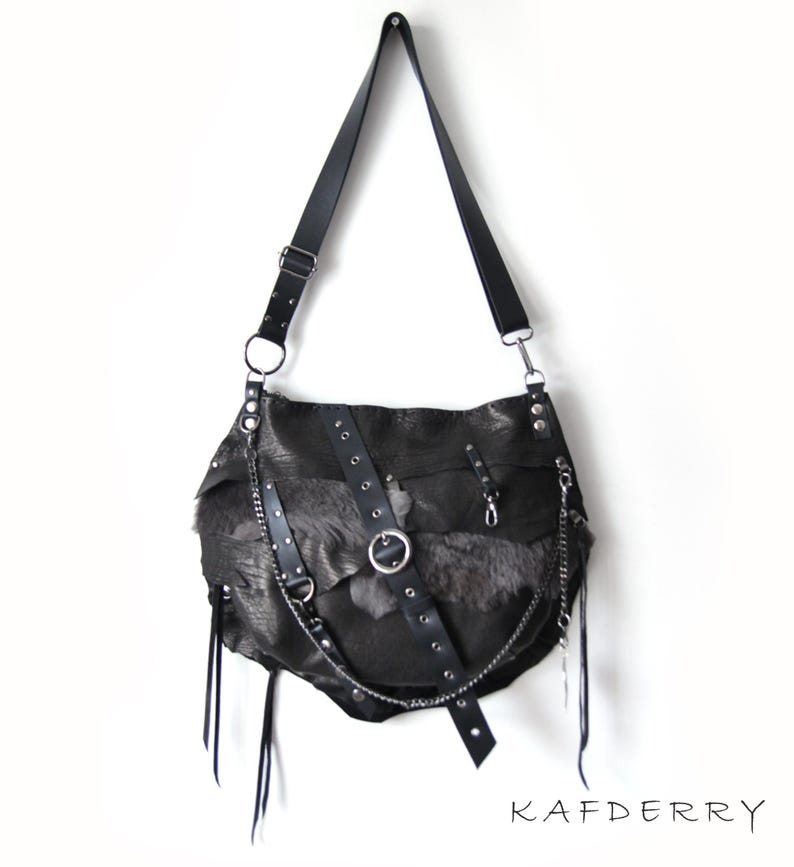 Puede incluir: Bolso de hombro de cuero negro con un dise&ntilde;o &uacute;nico. El bolso presenta una combinaci&oacute;n de texturas, incluyendo cuero, piel y cadenas met&aacute;licas. Tiene una correa larga y ajustable y correas decorativas de cuero con herrajes met&aacute;licos. El bolso est&aacute; suspendido sobre un fondo blanco.