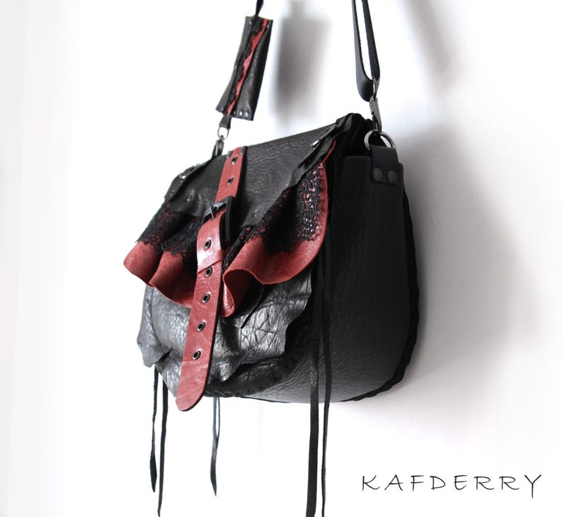 K&ouml;nnte beinhalten: Schwarze Leder-Umh&auml;ngetasche mit roten Akzenten. Die Tasche hat eine strukturierte Oberfl&auml;che, eine Klappe mit rotem Leder und Spitzentrimm, sowie einen roten Lederriemen mit Schnallen. Sie hat einen langen Schulterriemen und Lederkordeln.