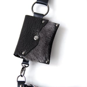 Puede incluir: Peque&ntilde;o bolso de cuero negro con solapa, adornado con piel gris y remaches plateados. El bolso est&aacute; sujeto a una correa de cuero negro con un anillo met&aacute;lico y un cierre plateado, sobre fondo blanco.