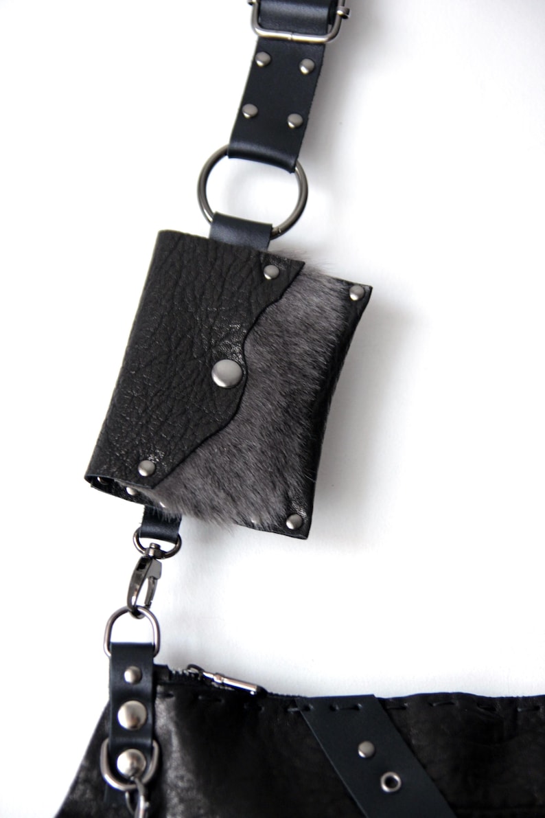 Puede incluir: Peque&ntilde;o bolso de cuero negro con solapa, adornado con piel gris y remaches plateados. El bolso est&aacute; sujeto a una correa de cuero negro con un anillo met&aacute;lico y un cierre plateado, sobre fondo blanco.