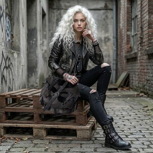 May include: A person wearing a black leather jacket, ripped black jeans, and black boots with buckles. A large, dark brown leather bag with buckles is slung over the shoulder. The person is sitting on a wooden pallet.