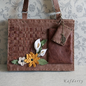May include: A brown leather handbag featuring a woven panel, a small attached pouch with a metal leaf charm, and decorative leather flowers in white, yellow, and green. The bag has visible stitching, metal rivets, and the text Kafderry in the corner.