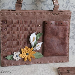 May include: A brown rectangular handbag featuring a woven leather panel, a smooth leather pocket with metal grommets, and decorative leather flowers in white, yellow, and green. The bag has visible stitching along the edges and metal rivets.