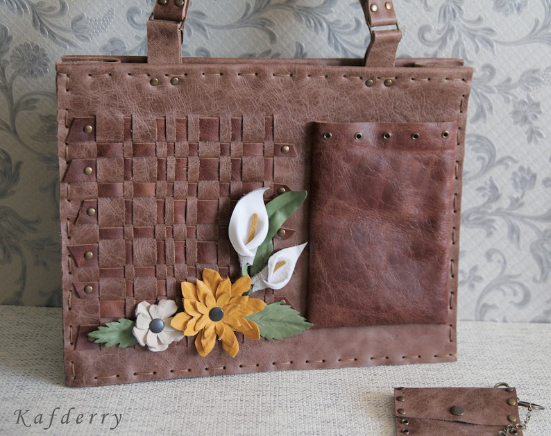 May include: A brown rectangular handbag featuring a woven leather panel, a smooth leather pocket with metal grommets, and decorative leather flowers in white, yellow, and green. The bag has visible stitching along the edges and metal rivets.
