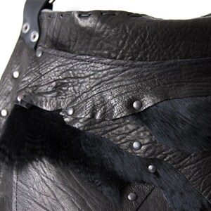 May include: Close-up of a black leather bag with a textured surface and silver rivets. The bag features a section of black fur and a stitched leather strap. The design is rugged and detailed.