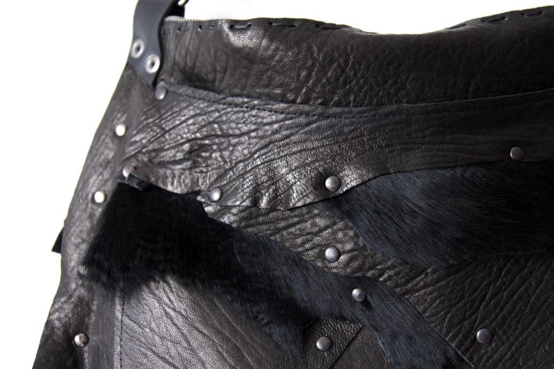 May include: Close-up of a black leather bag with a textured surface and silver rivets. The bag features a section of black fur and a stitched leather strap. The design is rugged and detailed.