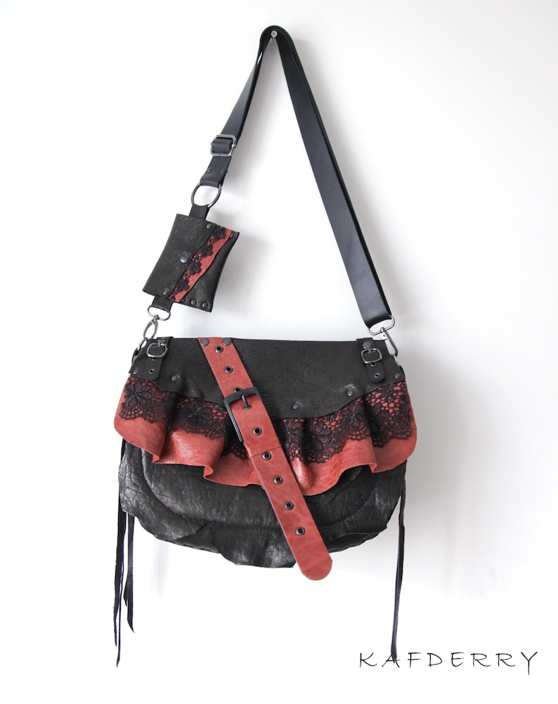 K&ouml;nnte beinhalten: Schwarze Leder-Umh&auml;ngetasche mit roter Leder- und schwarzer Spitzenr&uuml;sche. Die Tasche hat einen langen schwarzen Riemen und eine passende kleine Tasche. Ein roter Lederg&uuml;rtel mit Schnalle verl&auml;uft &uuml;ber die Vorderseite.