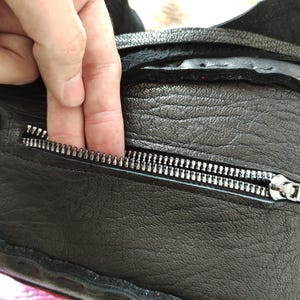 K&ouml;nnte beinhalten: Nahaufnahme einer schwarzen Ledertasche mit silbernem Rei&szlig;verschluss. Die Tasche hat eine strukturierte Oberfl&auml;che und sichtbare N&auml;hte. Eine Hand greift in die Rei&szlig;verschlusstasche und zeigt die Funktionalit&auml;t und das Design der Tasche.