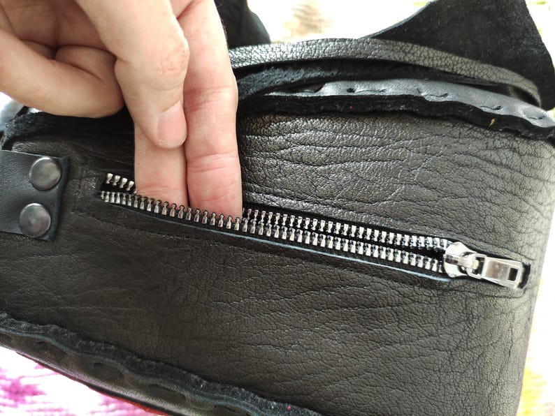 K&ouml;nnte beinhalten: Nahaufnahme einer schwarzen Ledertasche mit silbernem Rei&szlig;verschluss. Die Tasche hat eine strukturierte Oberfl&auml;che und sichtbare N&auml;hte. Eine Hand greift in die Rei&szlig;verschlusstasche und zeigt die Funktionalit&auml;t und das Design der Tasche.
