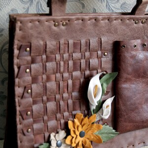 May include: A brown leather tote bag featuring a woven front panel, brass rivets, and decorative leather flower appliqu&eacute;s in white, yellow, and green. The bag includes a side pocket and two top handles. The word Kafderry is printed at the bottom.