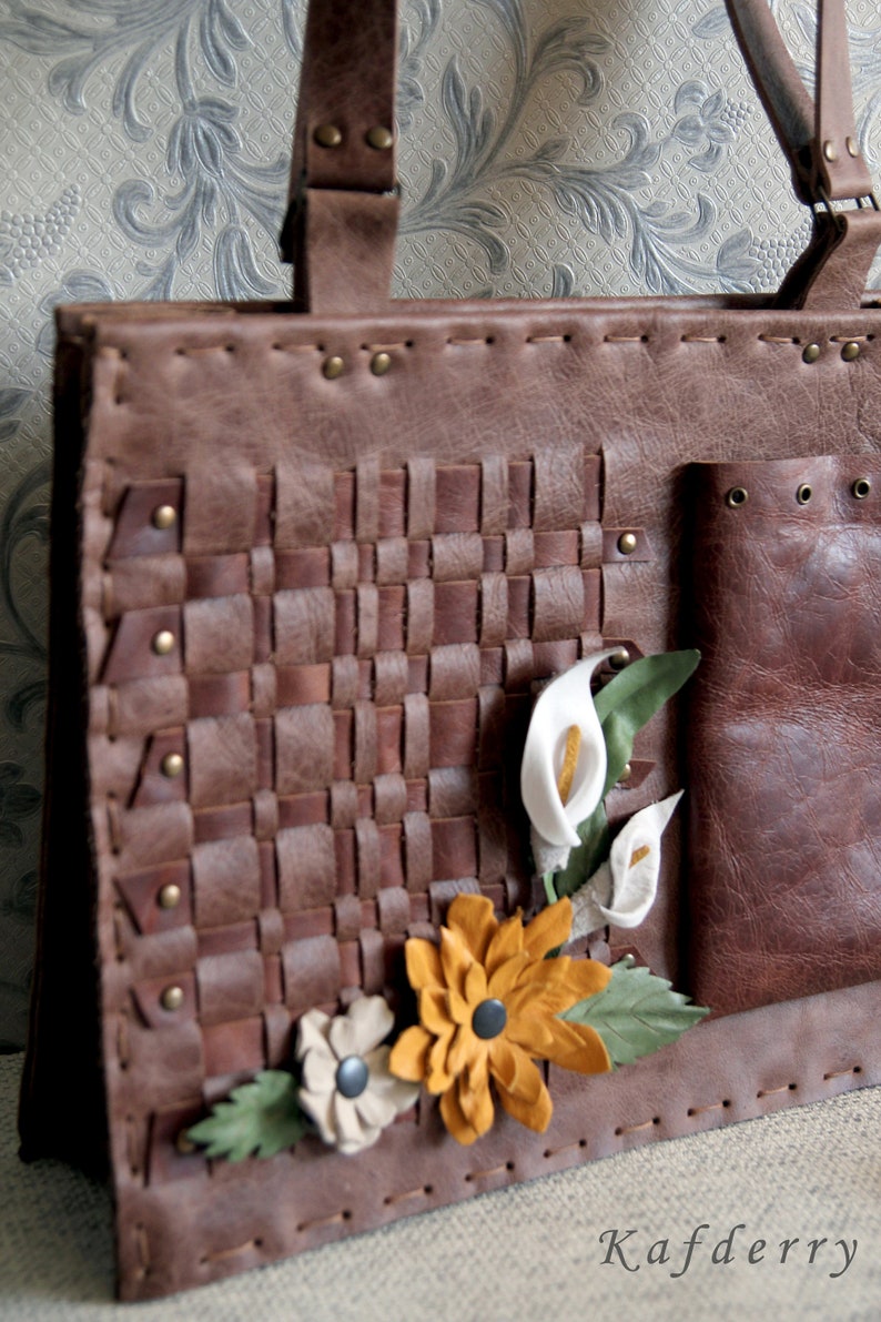 May include: A brown leather tote bag featuring a woven front panel, brass rivets, and decorative leather flower appliqu&eacute;s in white, yellow, and green. The bag includes a side pocket and two top handles. The word Kafderry is printed at the bottom.