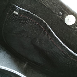 K&ouml;nnte beinhalten: Nahaufnahme des Innenraums einer schwarzen Ledertasche. Die Tasche hat eine strukturierte Tasche mit einer rot gen&auml;hten Naht, einen Metallverschluss und einen Magnetverschluss. Das Leder hat einen dezenten Glanz.