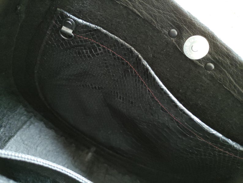 K&ouml;nnte beinhalten: Nahaufnahme des Innenraums einer schwarzen Ledertasche. Die Tasche hat eine strukturierte Tasche mit einer rot gen&auml;hten Naht, einen Metallverschluss und einen Magnetverschluss. Das Leder hat einen dezenten Glanz.