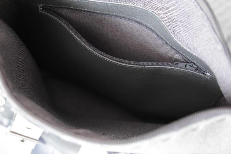 May include: Close-up of a grey handbag interior with a zipped pocket. The bag's interior is a lighter grey fabric, and the pocket is a darker grey material. The zip is silver.