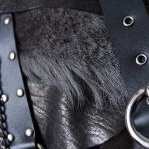 Puede incluir: Primer plano de una prenda de cuero negro con detalles de cadena y hebilla plateadas. El dise&ntilde;o incorpora una tira de piel sint&eacute;tica gris oscuro y una correa de cuero negro con remaches y ojales plateados. La est&eacute;tica general es oscura y texturizada.