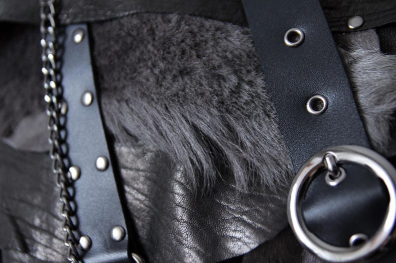 Puede incluir: Primer plano de una prenda de cuero negro con detalles de cadena y hebilla plateadas. El dise&ntilde;o incorpora una tira de piel sint&eacute;tica gris oscuro y una correa de cuero negro con remaches y ojales plateados. La est&eacute;tica general es oscura y texturizada.