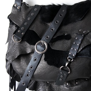 May include: Black leather shoulder bag with layered design and fur accents. Features multiple straps with metal hardware, including rivets and a large silver ring. The bag has a fringe detail.
