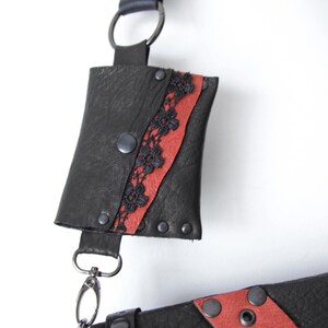 K&ouml;nnte beinhalten: Schwarze Leder-Umh&auml;ngetasche mit rotem Streifen und schwarzen Spitzendetails. Die Tasche hat einen Druckknopfverschluss und Metallbeschl&auml;ge. Der Riemen ist verstellbar und hat eine kleine Tasche.