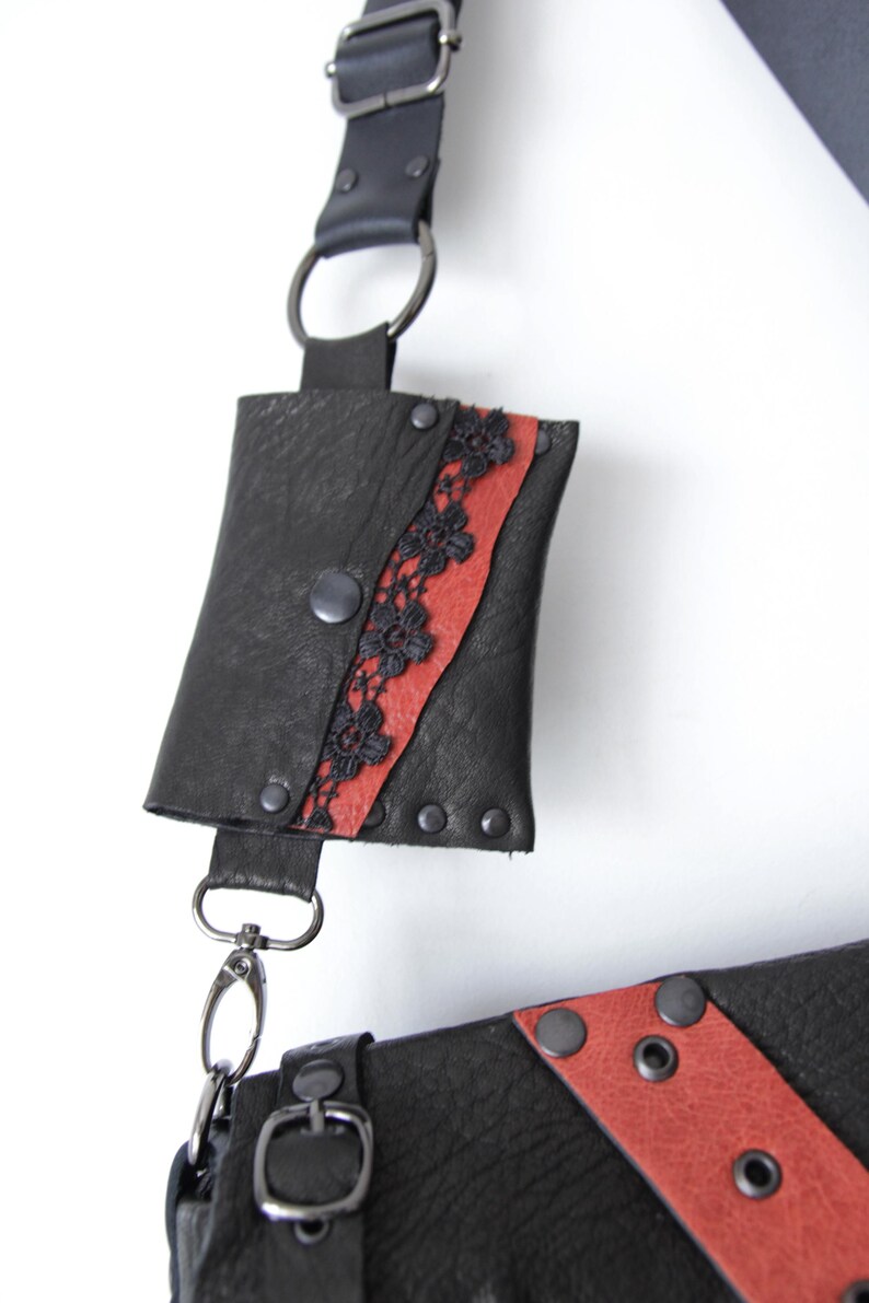 K&ouml;nnte beinhalten: Schwarze Leder-Umh&auml;ngetasche mit rotem Streifen und schwarzen Spitzendetails. Die Tasche hat einen Druckknopfverschluss und Metallbeschl&auml;ge. Der Riemen ist verstellbar und hat eine kleine Tasche.