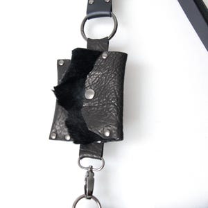 May include: Black leather pouch with a flap, secured by a silver snap, attached to a black strap with a silver clasp. The pouch has a textured surface and a soft, black interior. The strap is connected to a larger bag.