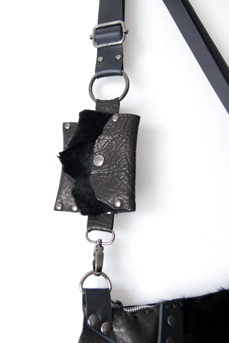 May include: Black leather pouch with a flap, secured by a silver snap, attached to a black strap with a silver clasp. The pouch has a textured surface and a soft, black interior. The strap is connected to a larger bag.