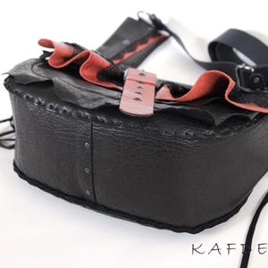 K&ouml;nnte beinhalten: Schwarze Leder-Umh&auml;ngetasche mit rotem und schwarzem R&uuml;schenbesatz. Die Tasche hat einen rosafarbenen Schnallenverschluss und einen schwarzen Riemen. Sie ist mit Nieten und N&auml;hten versehen.