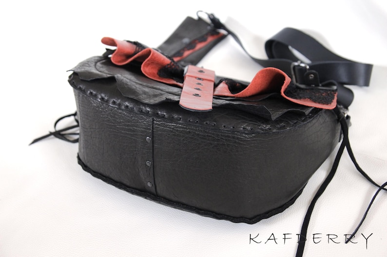 K&ouml;nnte beinhalten: Schwarze Leder-Umh&auml;ngetasche mit rotem und schwarzem R&uuml;schenbesatz. Die Tasche hat einen rosafarbenen Schnallenverschluss und einen schwarzen Riemen. Sie ist mit Nieten und N&auml;hten versehen.