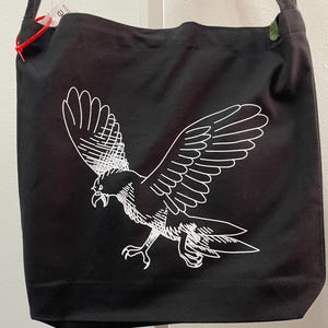May include: Black tote bag with a white graphic of a bird in flight. The bag has a shoulder strap and a rectangular shape. The bird design is detailed with white lines, and the bag appears to be made of canvas or a similar material.