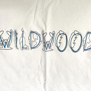 May include: White t-shirt with the word "WILDWOOD" in blue and white lettering. Each letter is designed with a unique illustration, including a seal, a cat, and other whimsical creatures.